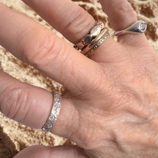 Sterling silver sandcast ring with flush-set cubic zirconias, worn on a hand resting gently over sand — the ring’s textured surface echoing the natural patterns beneath.