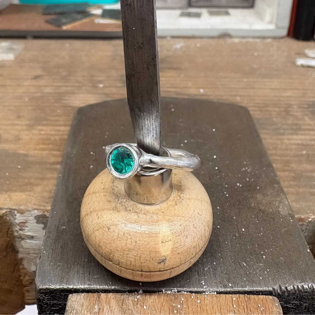 Cherish Ring in sterling silver, Size O, supported on a graver tool and viewed from a front top angle slightly to the right, showing the flush-set Biron emerald.
