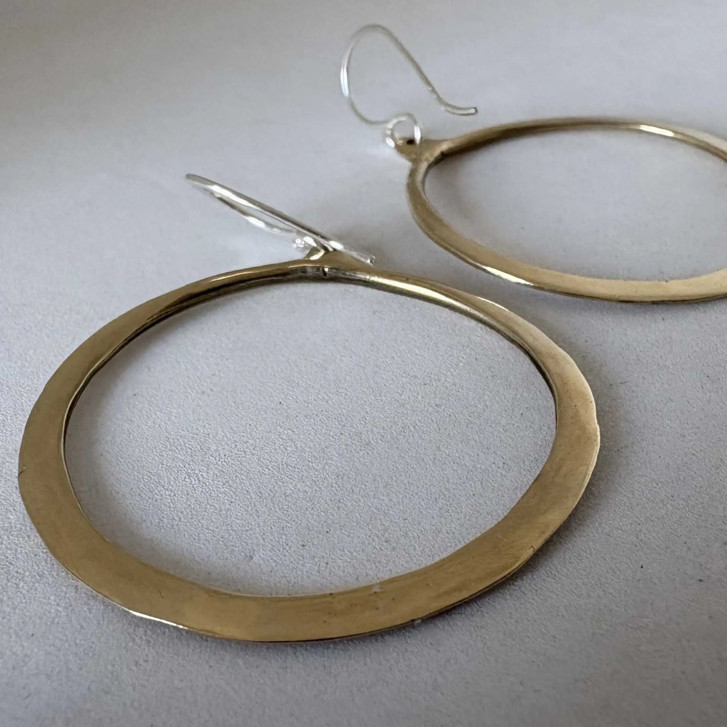 Close-up of Lunar Eclipse hoop earrings in polished golden bronze with sterling silver hooks, one hoop in forefront against light grey textured background