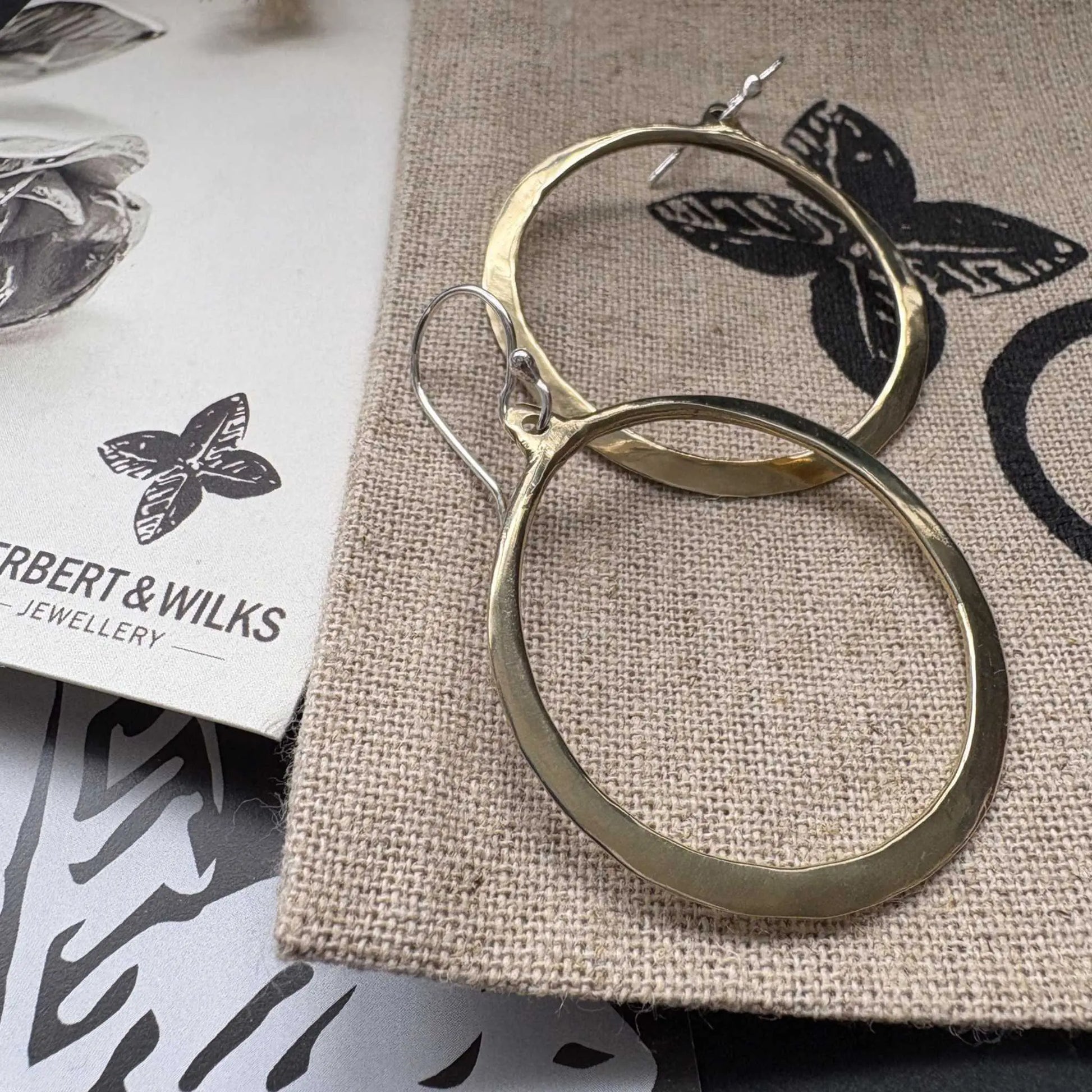 Close-up of Lunar Eclipse hoop earrings in polished golden bronze with sterling silver hooks, one hoop in forefront against light grey textured background