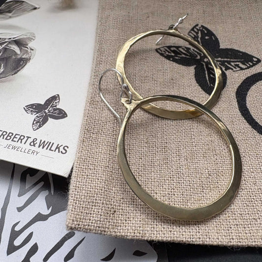 Close-up of Lunar Eclipse hoop earrings in polished golden bronze with sterling silver hooks, one hoop in forefront against light grey textured background