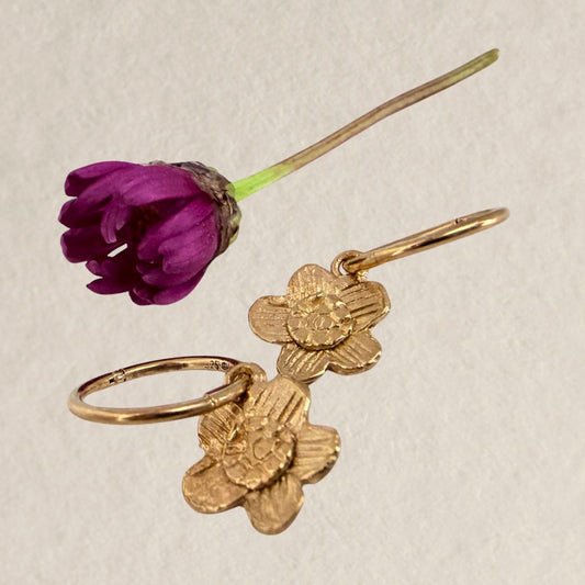 A pair of 18ct gold-plated alpine daisy charm sleeper earrings with textured petals and circular sleeper hoops, displayed on a light beige textured background next to a purple flower.