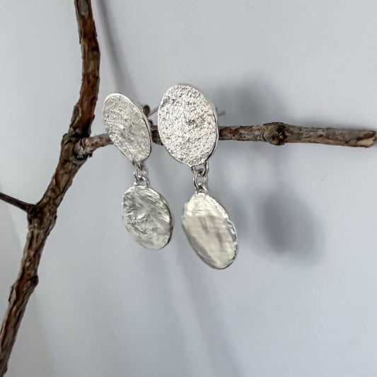 Artistic side-angle view of the sterling silver Sequin Disc Dangle Earrings hanging from a delicate branch, with soft shadows on the background wall highlighting their thin, lightweight profile.