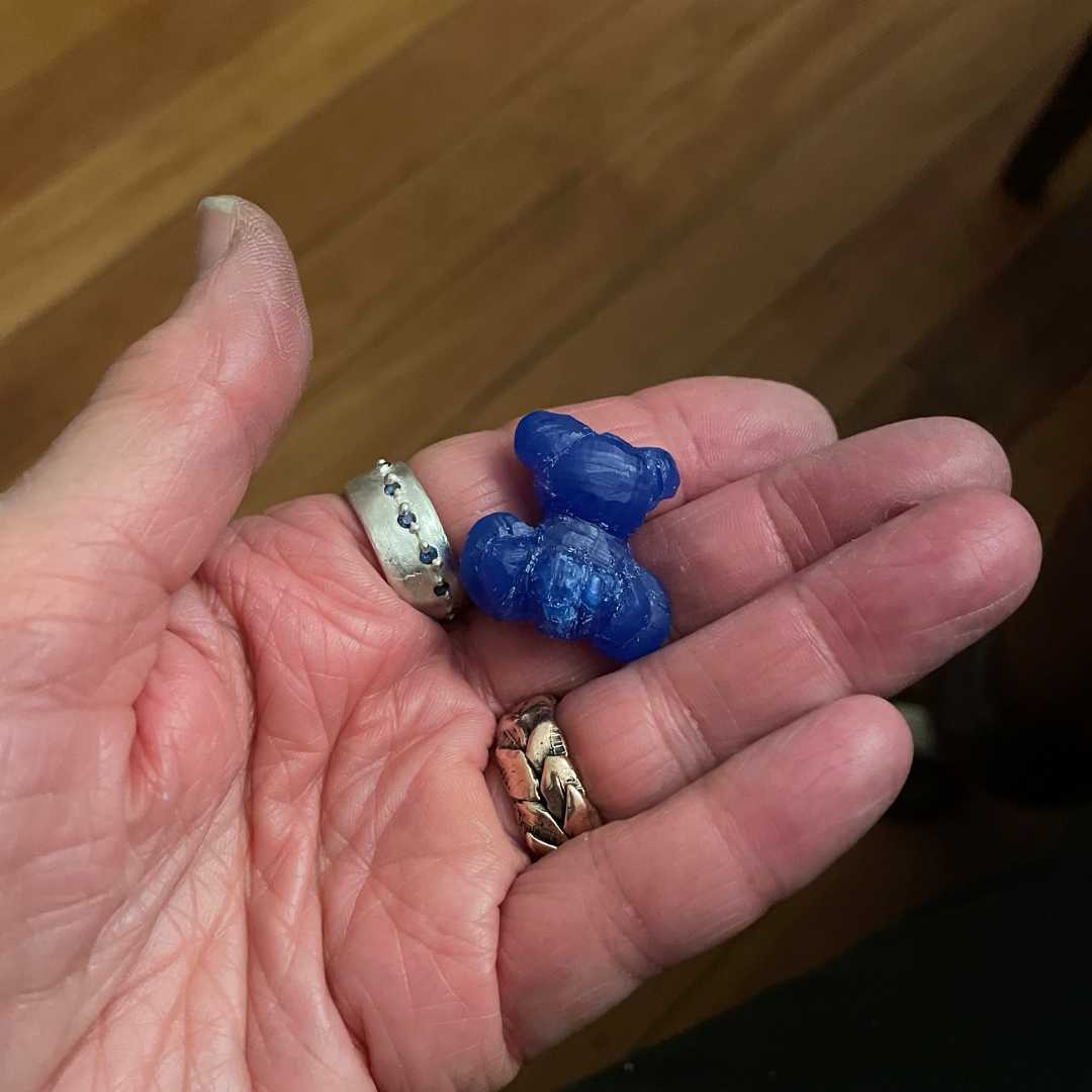 A work in process of a Blue wax teddy bear pendant being held in a hand over a wooden work bench