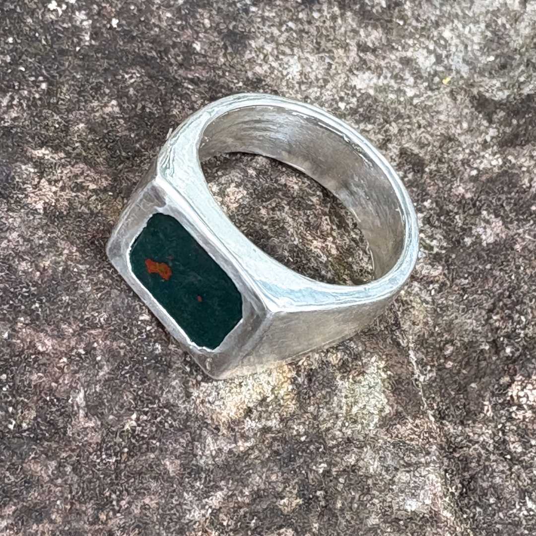 Top-down angled view from the right of the Rectangle Bloodstone Signet Ring resting on textured basalt rock in greys and whites, highlighting the green Bloodstone and hand-polished silver.