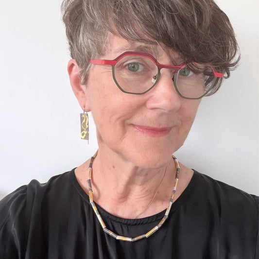 Woman with short hair wearing red and green glasses and a black dress, wearing wrapped fine silver and gold beaded necklace with matching gilded earrings