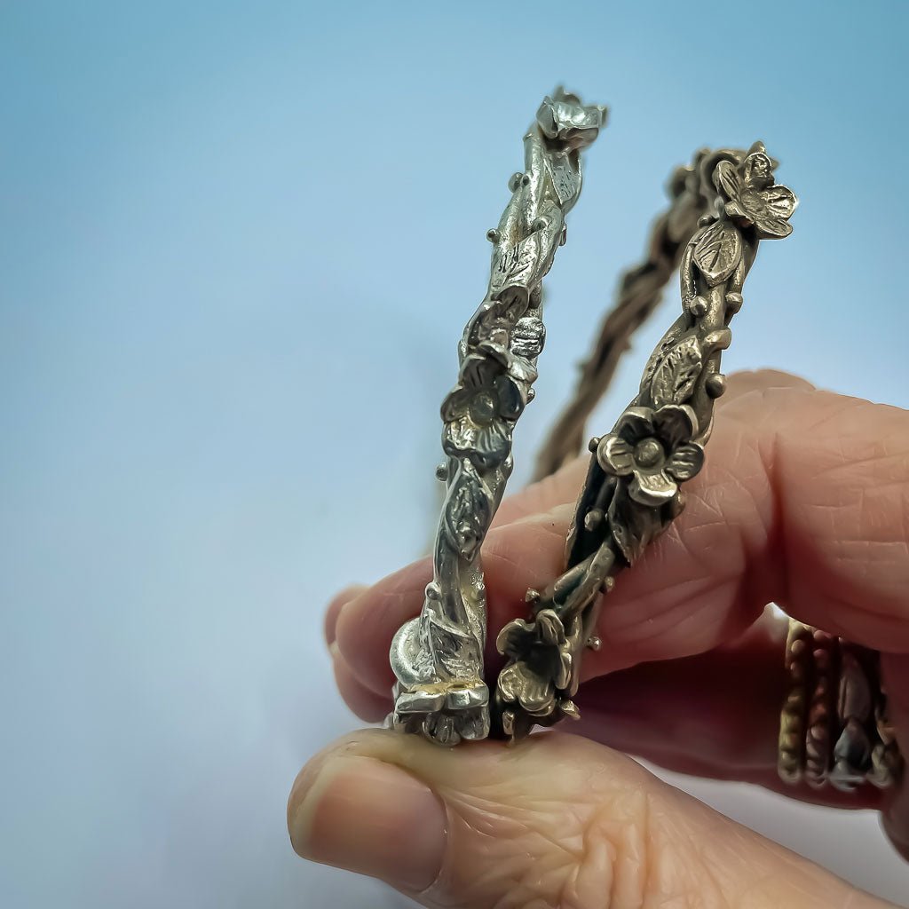 Sterling silver and bronze Interwoven Flora Bangles held up to show sculptural flower, leaf, and vine details against a blue background.
