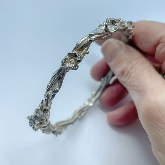 Sterling silver Interwoven Flora Bangle held up to show sculptural flower, leaf, and vine details against a light blue background.