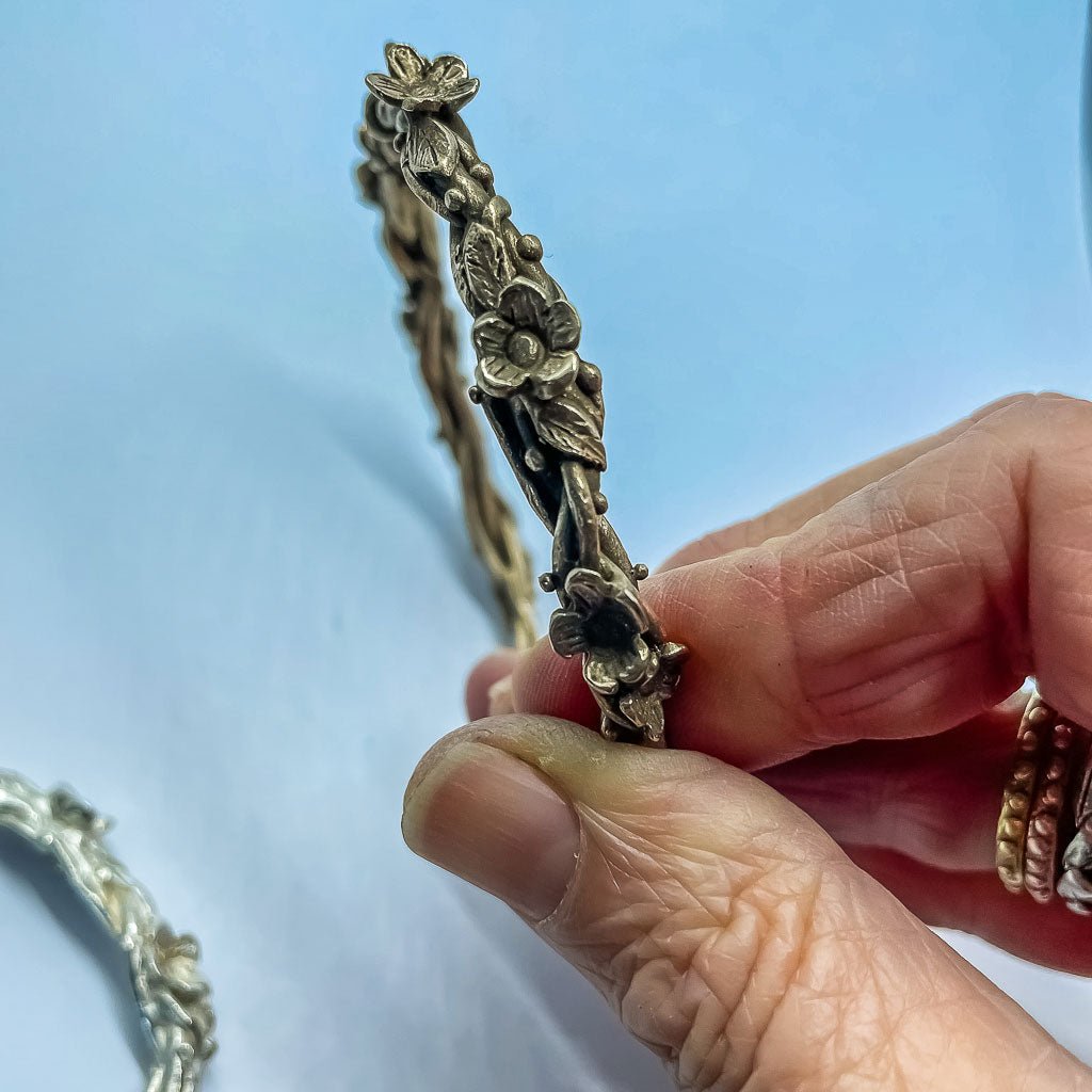 Bronze Interwoven Flora Bangle held up against a blue background, showing sculptural flower, leaf, and vine details.