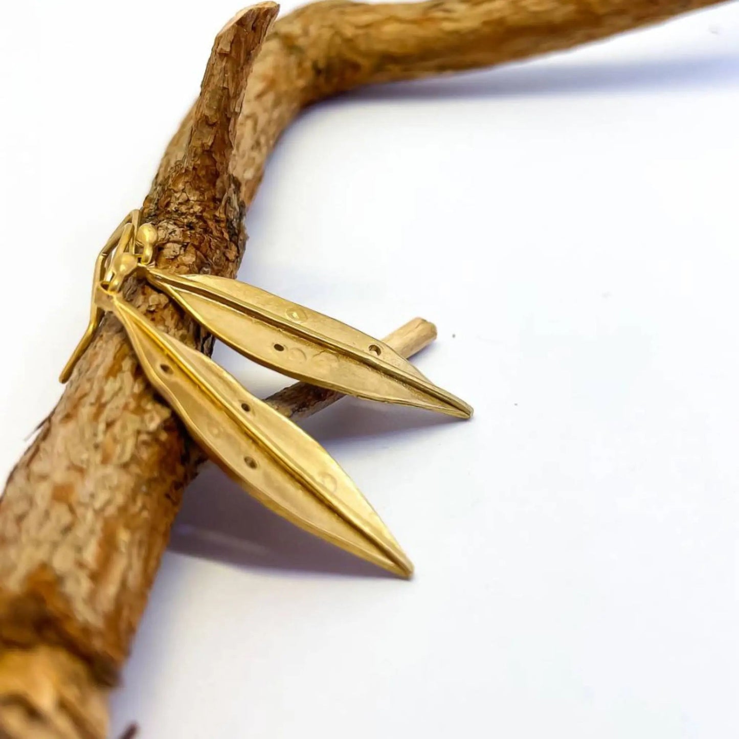 a side profile for the 18ct Gold Plate over Sterling Silver Pōhutukawa Leaf Pendant Earrings leaning against a branch with textured bark, The edge of the earring is visible