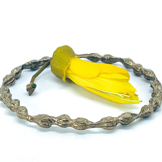 A bronze Kōwhai Pod bangle resting on a white surface, with a sculptural yellow kōwhai flower placed in the centre of the bangle.