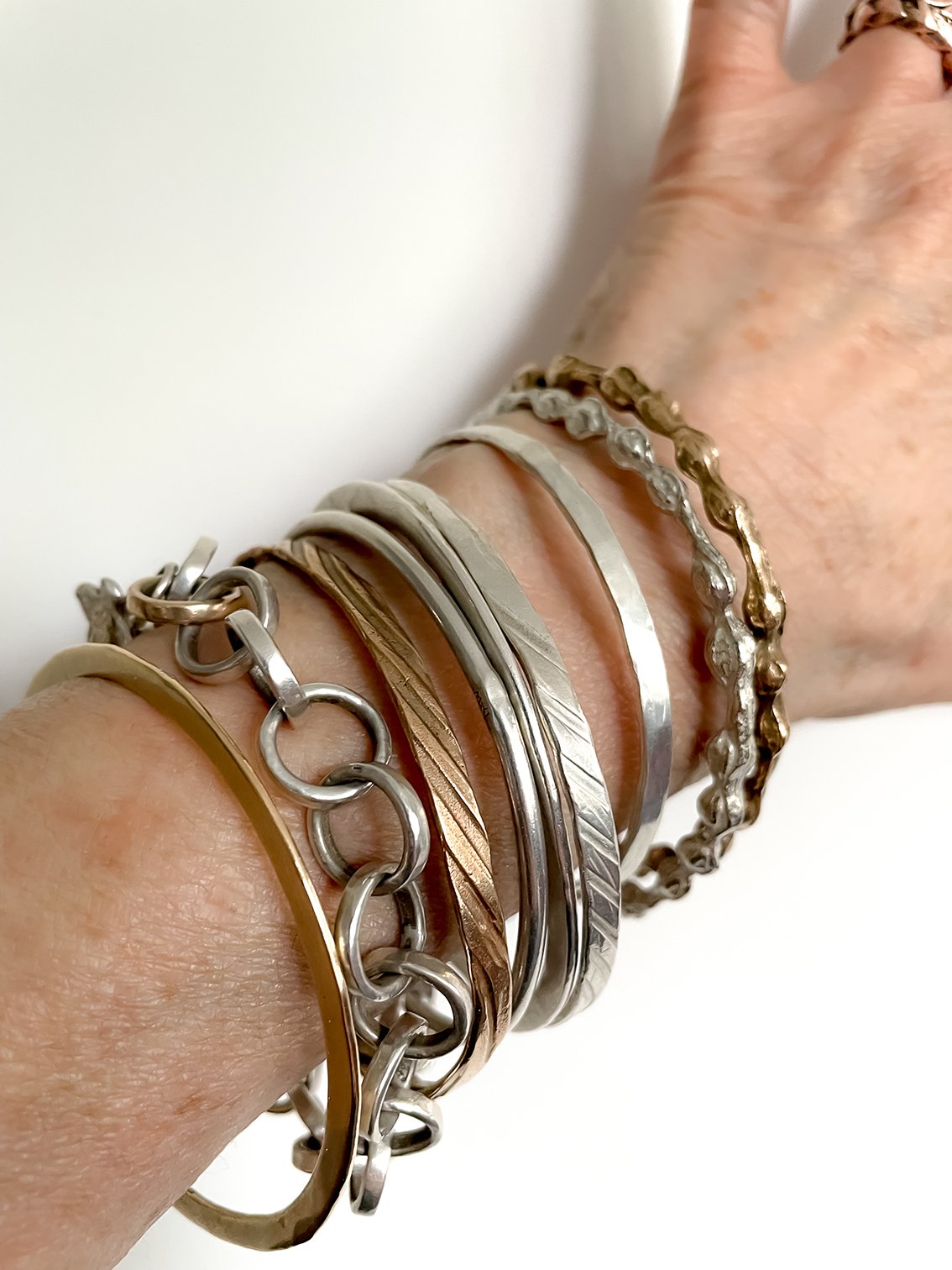 Kōwhai Pod Sterling Silver Bangle stacked with a mix of silver, gold, and bronze bangles and chains, showcasing its sculptural shape and versatility in a layered look.