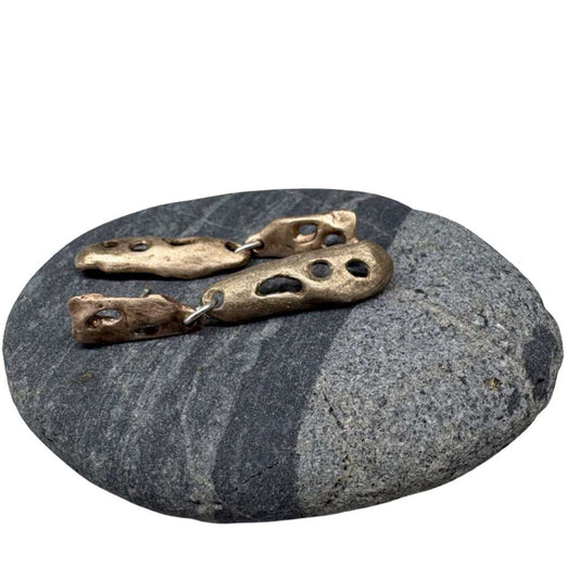 Bronze Perfora Curve Drop Earrings lying on smooth, striped river pebble, highlighting the warm metal tones and organic inspiration behind the design