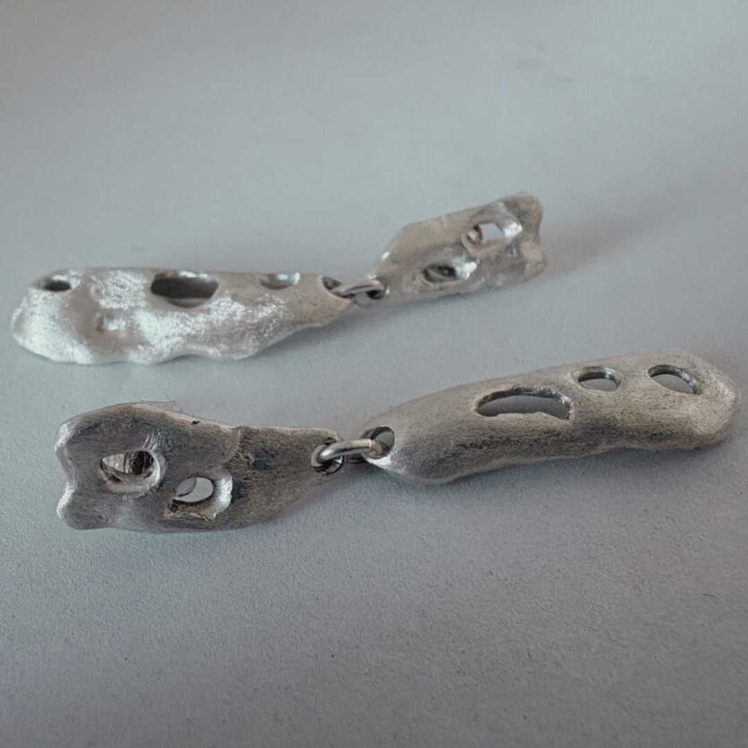 Artistic shot of the sterling silver Perfora Curve Drop Earrings lying on their sides against a grey surface, captured in soft, grainy focus to emphasise form and shadow.