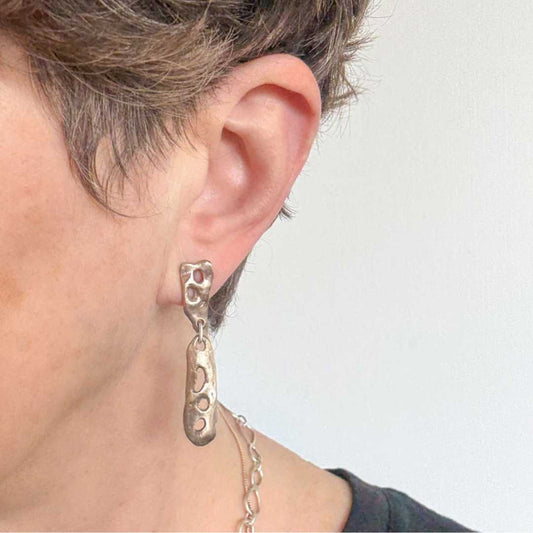 Close-up of the bronze Perfora Curve Drop Earrings worn by a woman with short hair, photographed against a light grey background to highlight the warm tones and sculptural curves.