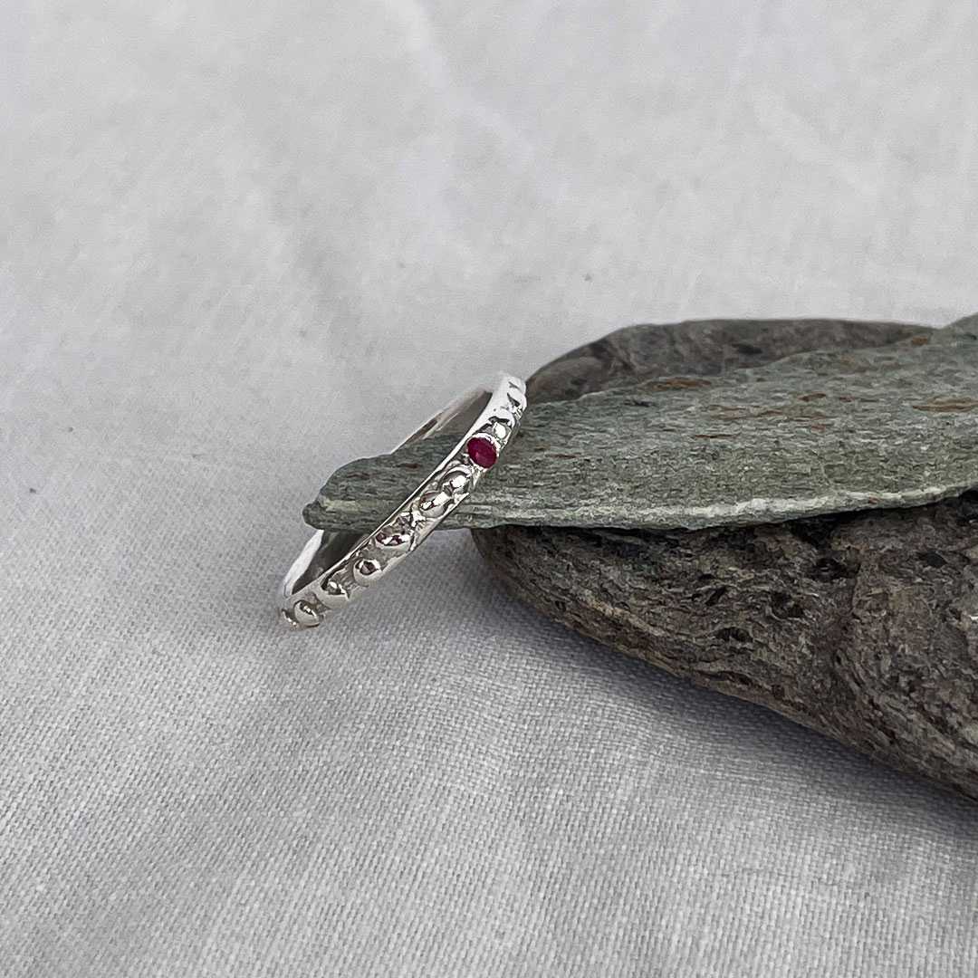 Fluid Ember Stacking Ring in sterling silver with a flush-set natural ruby, top-down view angled to the right, placed beside grey stones on a cream linen background.