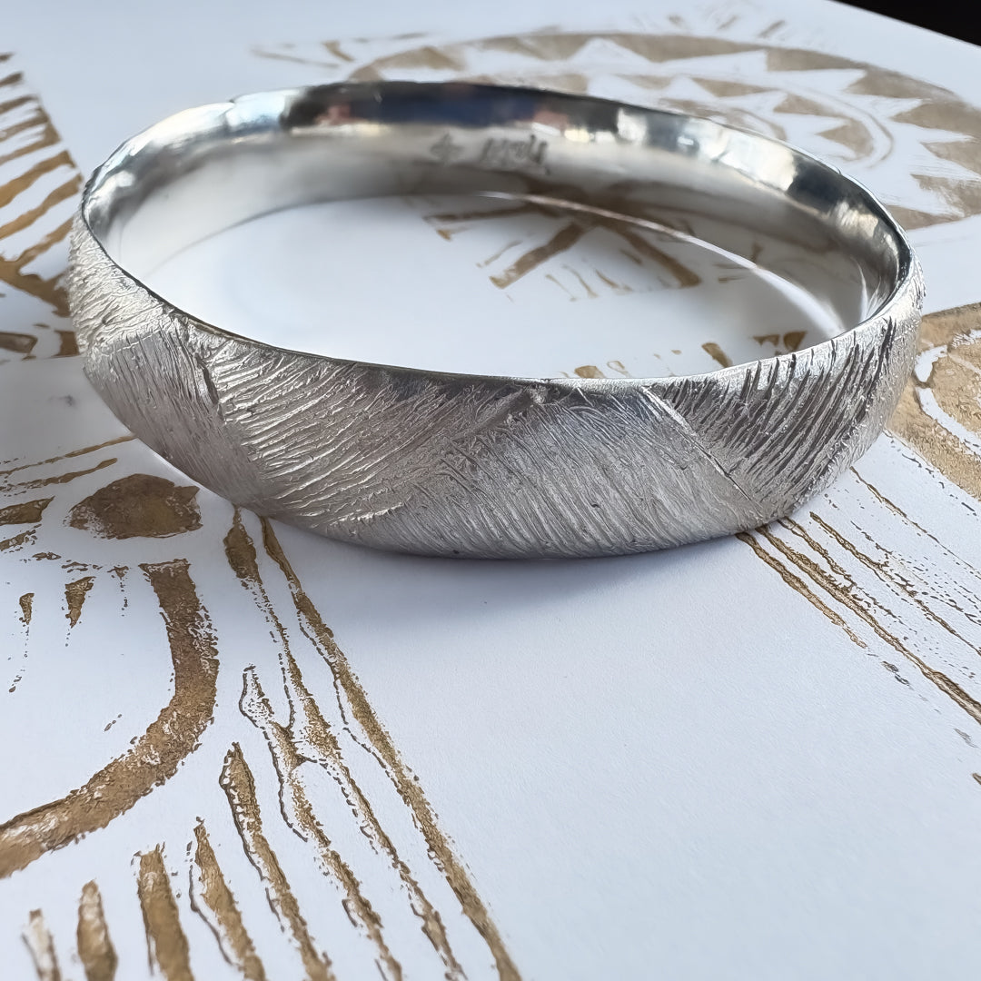 Strata Silver Bangle sitting on gold lino prints on white paper — showing how the woven surface texture echoes the printmaking lines that inspired HerbertandWilks’ design.