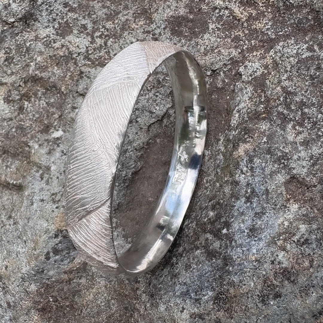 Top-down view of the Strata Silver Bangle on basalt rock showing engraved H&W and flower emblem inside the polished band with textured exterior surface.
