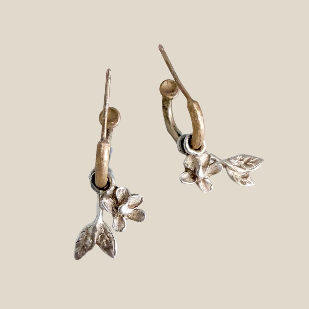 A pair of 9ct gold hoop earrings with with textured sterling silver leaf and flower charms, shown on a plain beige background.