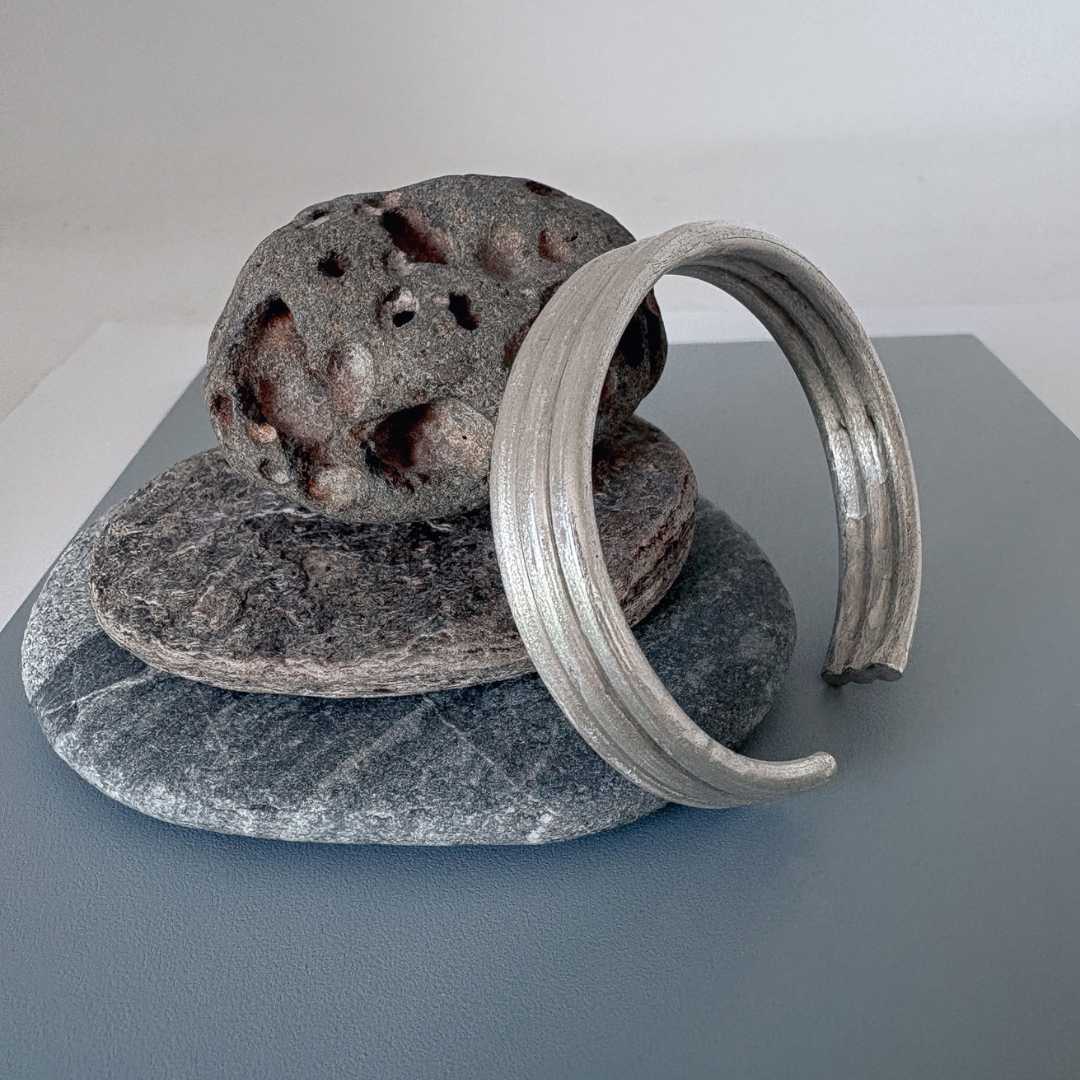 Three Line Cuff in sterling silver resting against a stack of three rocks on a steel-blue board with a light grey border, highlighting its sculptural, minimal design