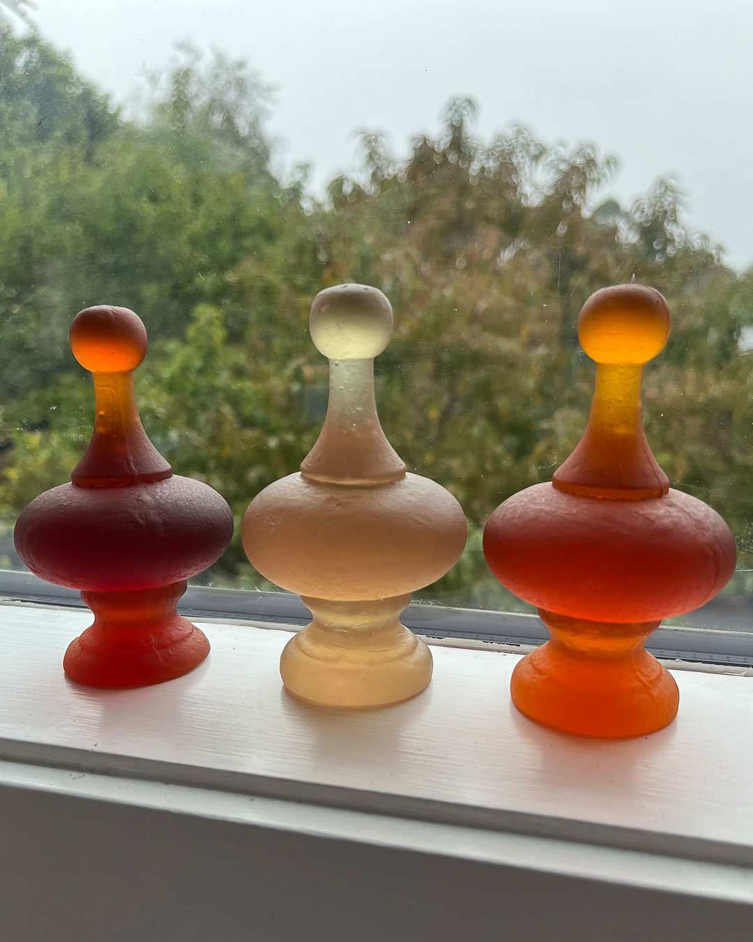 A group of three cast glass finials on a window sill in Dark Orange, Orange and Rhubarb. They form wonderful light-filled installations in groups.