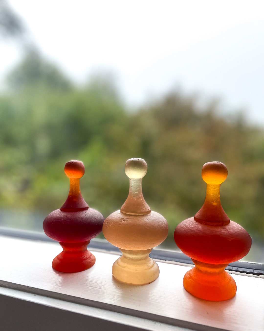 A group of three cast glass finials on a window sill in Dark Orange, Orange and Rhubarb. They form wonderful light-filled installations in groups.
