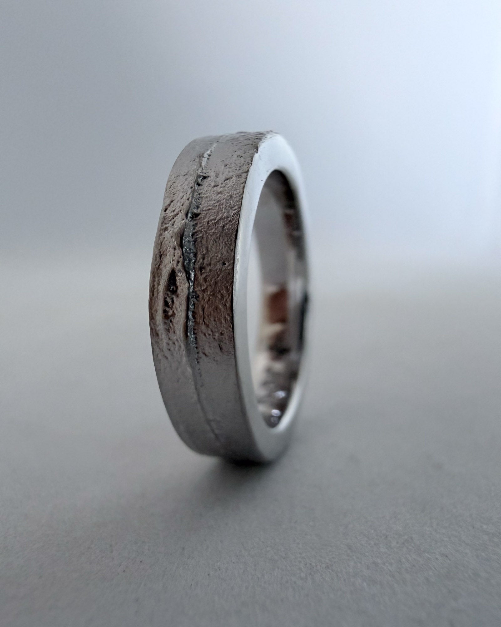 Three-quarter view displaying both the uniquely textured flat front face and the smooth side profile of the rhodium-plated sandcast band on a blue-grey background.