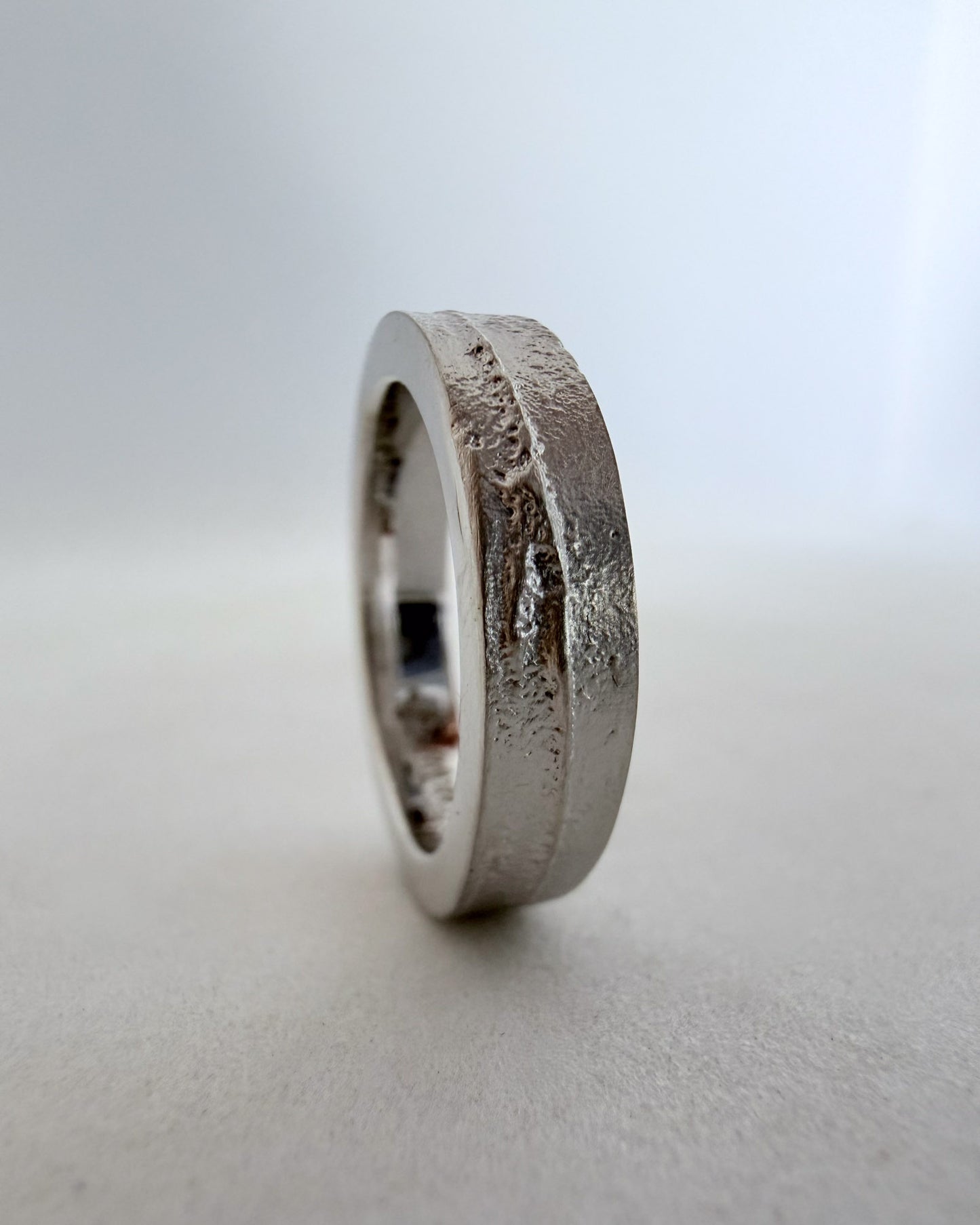 Close-up view of the front face of a 4mm rhodium-plated white gold sandcast ring, highlighting the raw, organic texture against a blue-grey background.