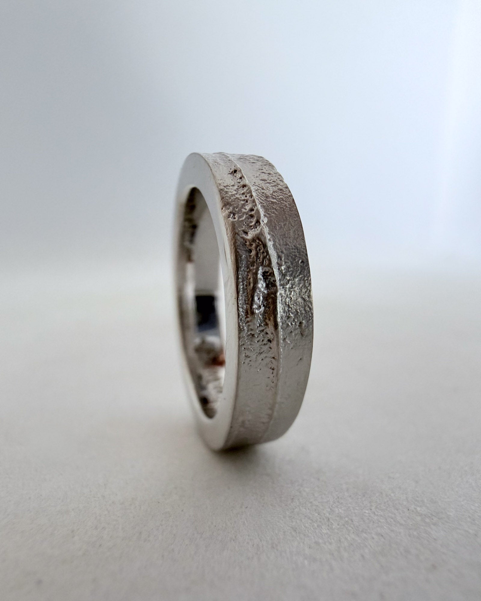Close-up view of the front face of a 4mm rhodium-plated white gold sandcast ring, highlighting the raw, organic texture against a blue-grey background.