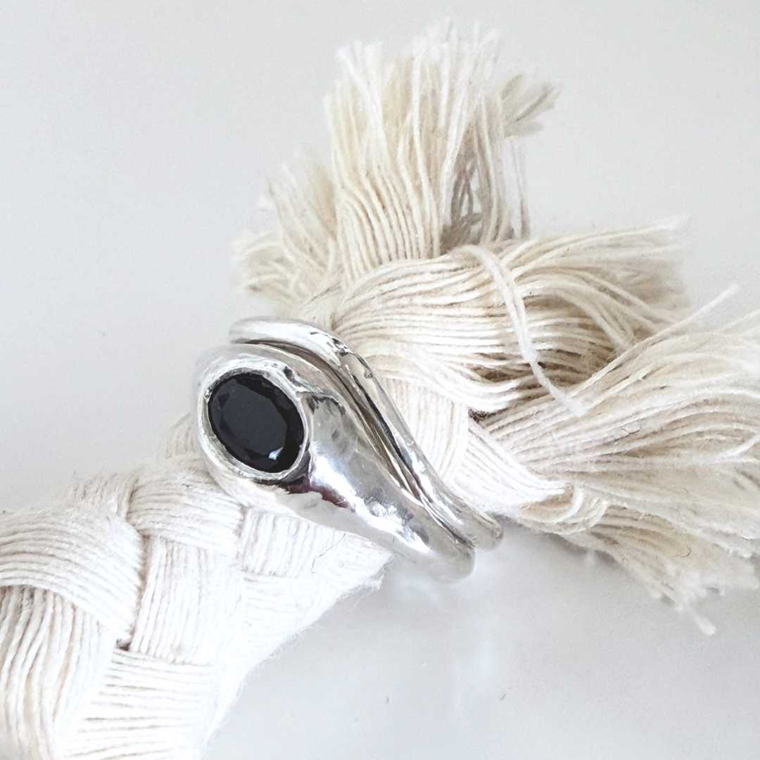 The Kāmāhi sterling silver and black sapphire signet ring nestled alongside the plain Sylvan curved band. Photographed from a slight front-right angle looking down on a cream braided cord, showcasing how the two rings stack together as an alternative bridal set.