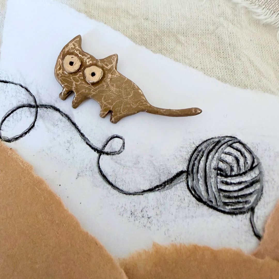 Bronze Cat Pin beside a hand-drawn ball of wool on textured paper and fabric
