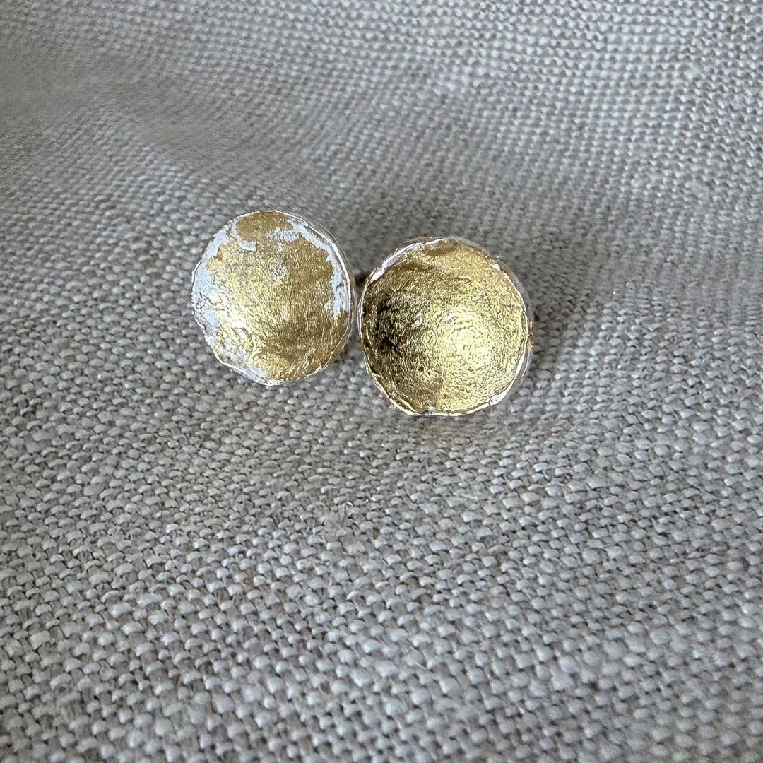 The Medium sized wrapped concave stud earrings in fine silver and 24 ct gold leaf lying on light coloured textured linen