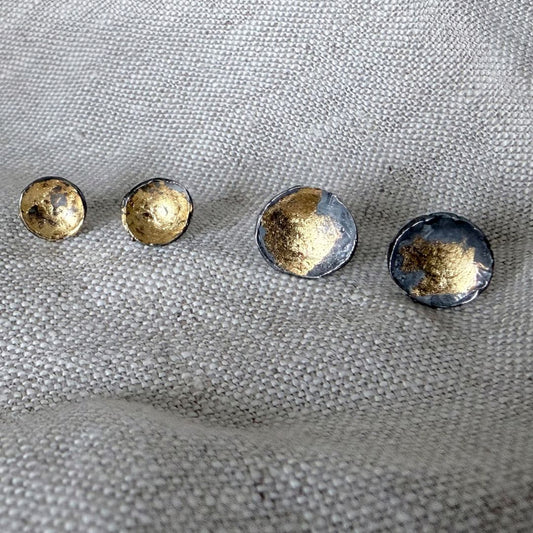 The Small and Medium sized wrapped concave stud earrings in oxidised fine silver and 24 ct gold leaf sitting on linen material.