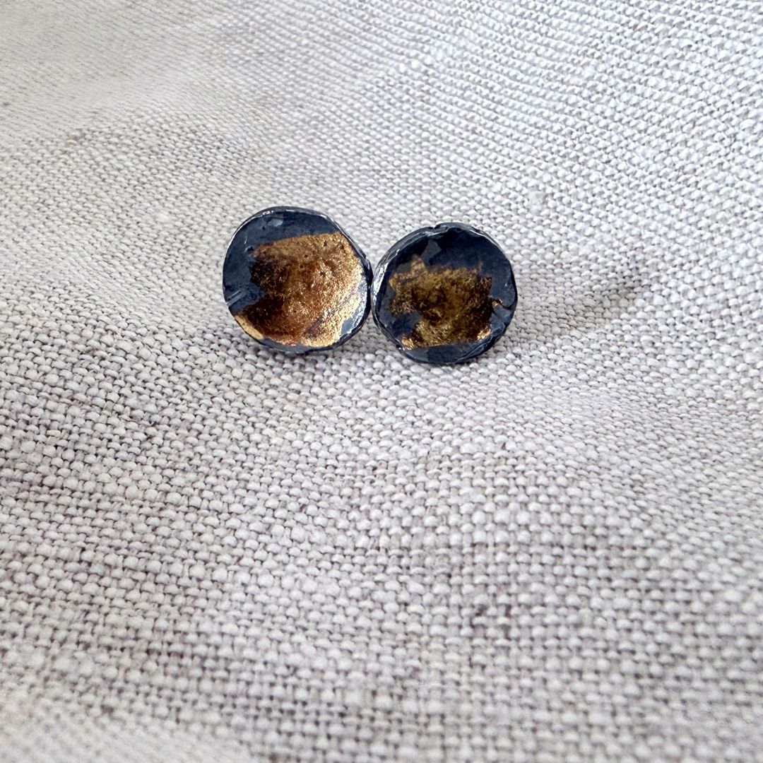 The Medium sized wrapped concave stud earrings in oxidised fine silver and 24 ct gold leaf lying on light coloured textured linen
