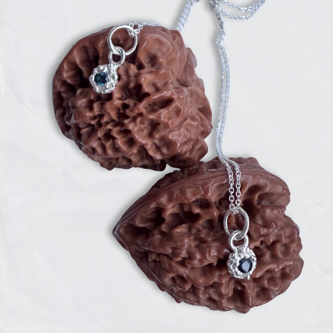 Macro view of two organic, textured sterling silver Fluid Dewdrop pendants with cast-in-place blue sapphires resting on dark brown seed pods.