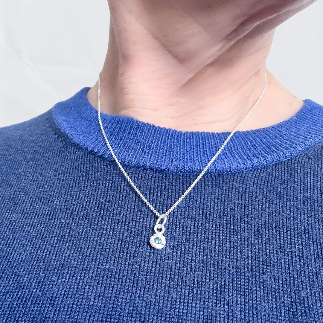 Close-up of a silver Dewdrop sapphire pendant resting below the collarbone against a blue ribbed sweater, demonstrating necklace length and fit.