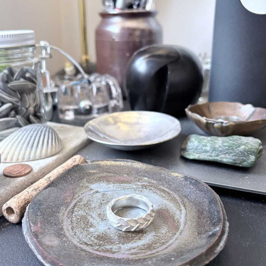 Hand-carved sterling silver laurel leaf wedding band displayed in ceramic pottery bowl on wooden desk, botanical jewellery lifestyle photography