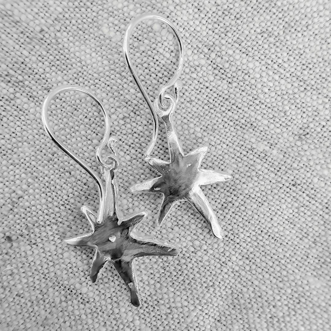 Back view of sterling silver Faerie Star dangle earrings resting on textured linen, showing the handmade organic star shapes and ear hooks.