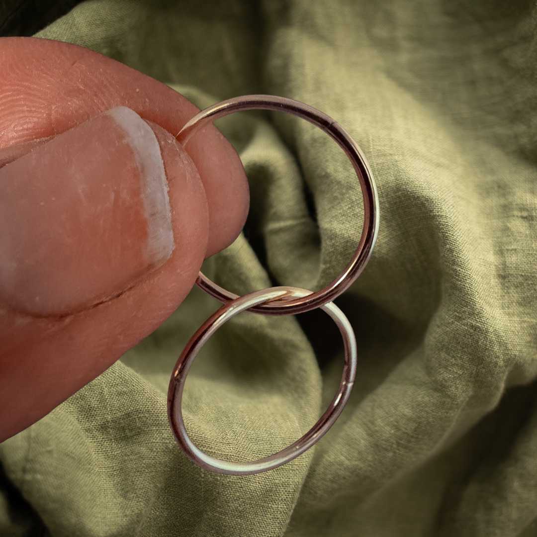 Pair of 16mm 9ct gold sleeper earrings interlinked together with the top hoop held between fingers against a textured green linen fabric background, showcasing the slightly bolder standalone scale and rich yellow gold color.