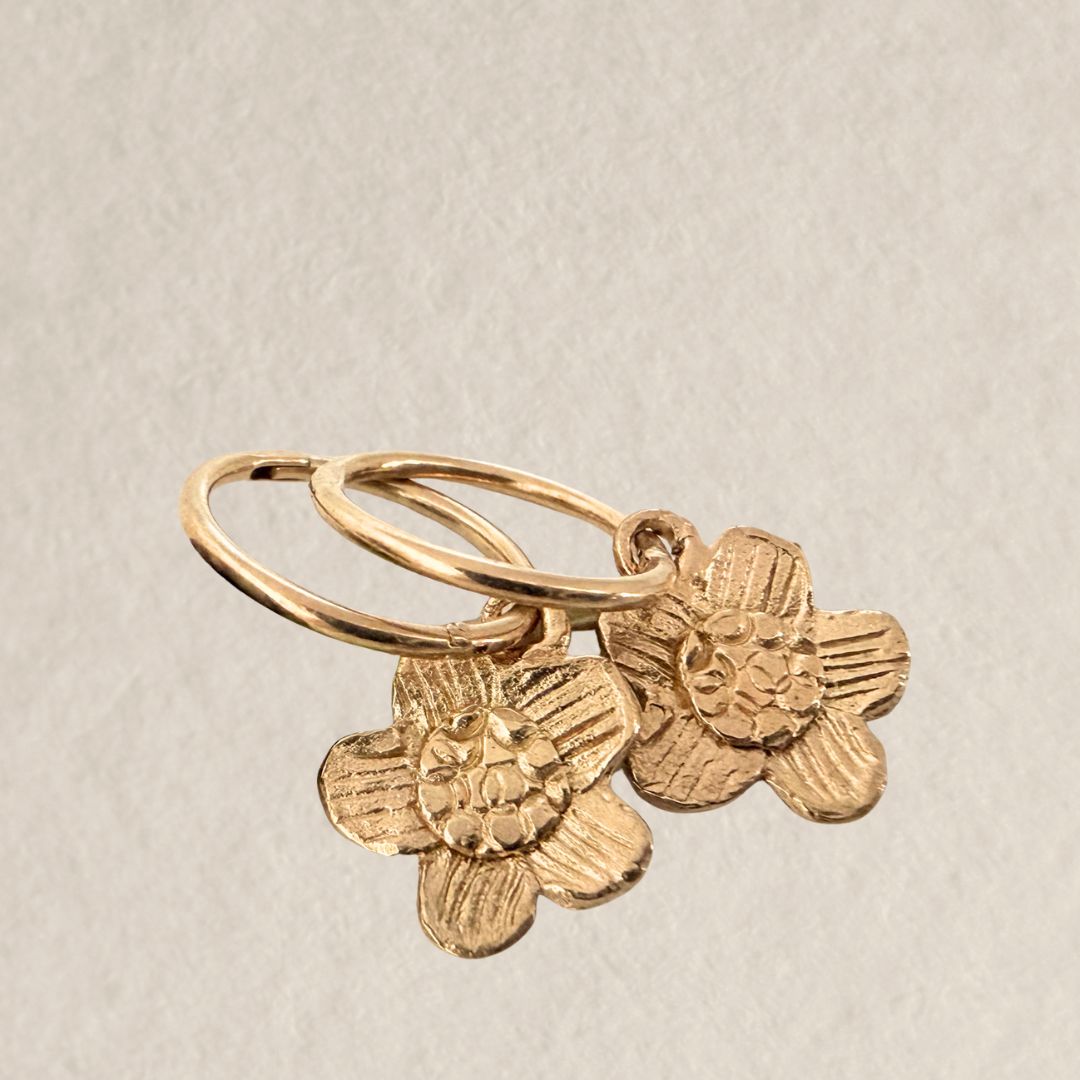 Pair of 18ct gold-plated alpine daisy charm sleeper earrings lying flat on a beige textured stone background.