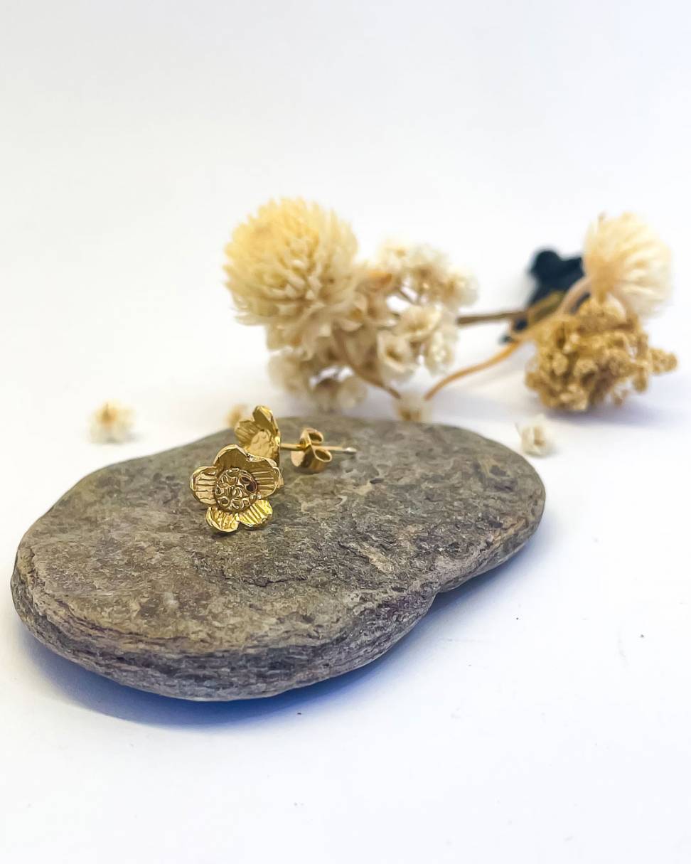 a pair of Golden Daisy Flower Stud Earrings sitting on a rock with flowers behind