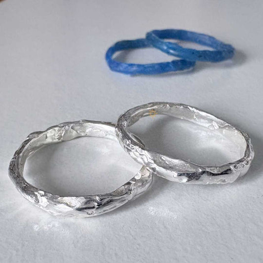 Two polished sterling silver stacking rings on light grey surface with blue wax ring models behind - wax build-up jewellery making workshop