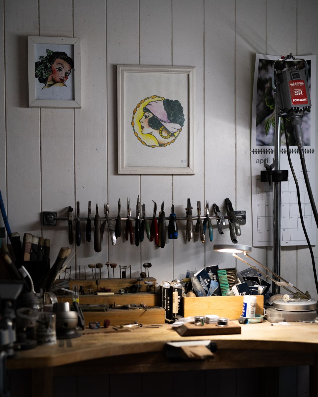 Workshop with nultiple pliers hanging on a wall rack and jewellers work bench with various items.