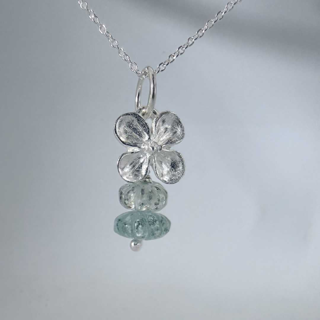 Close-up of hydrangea and aquamarine necklace in sterling silver hanging against a light grey wall, showing soft shadows and handcrafted texture