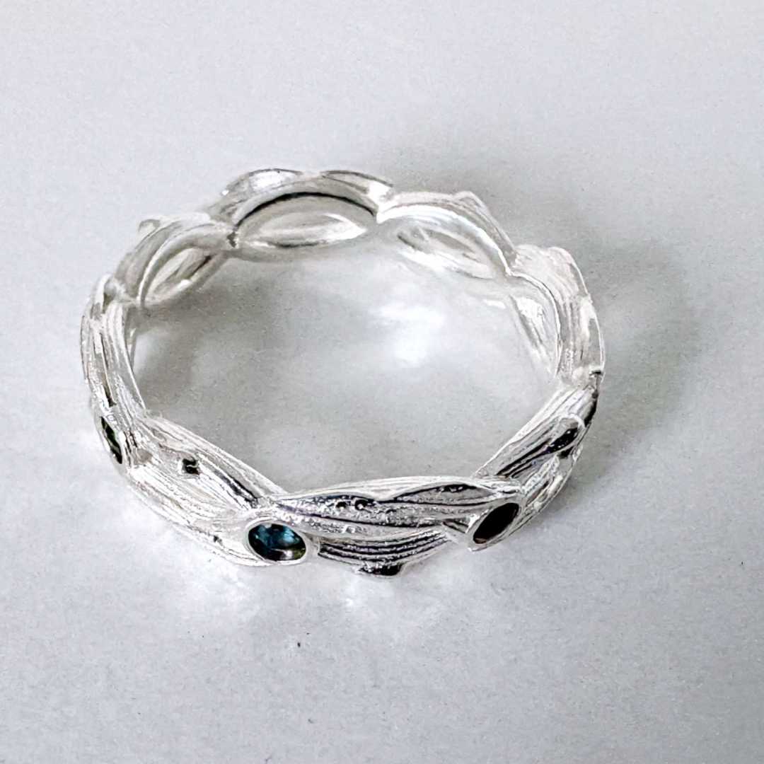  Interwoven sapphire ring lying flat on grey surface, overhead view showing botanical leaf detail and bark texture across band