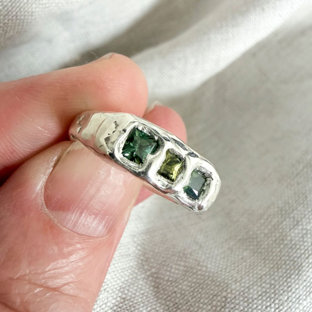 Close-up of the Kākāriki Signet Ring held between fingers, showing three princess-cut green sapphires and the irregular sculptural silver setting.