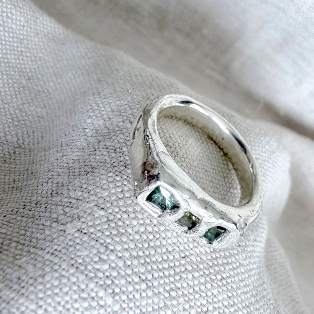 Angled view of the Kākāriki Signet Ring on natural linen, highlighting the sculptural silver contours and three green sapphires.