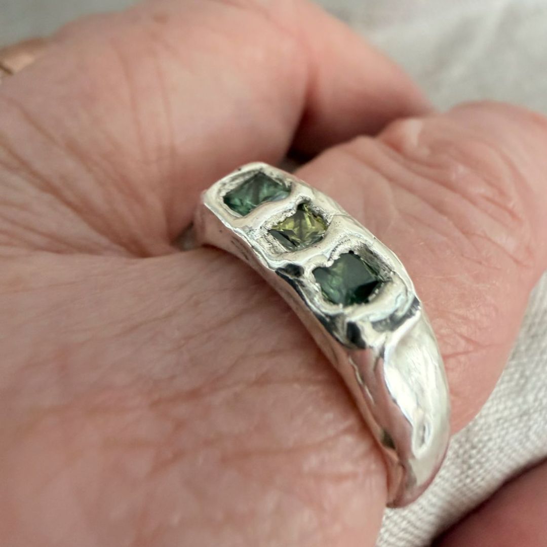 Kākāriki sterling silver signet ring worn on a finger, showing its softly polished organic band and three green princess-cut sapphires.