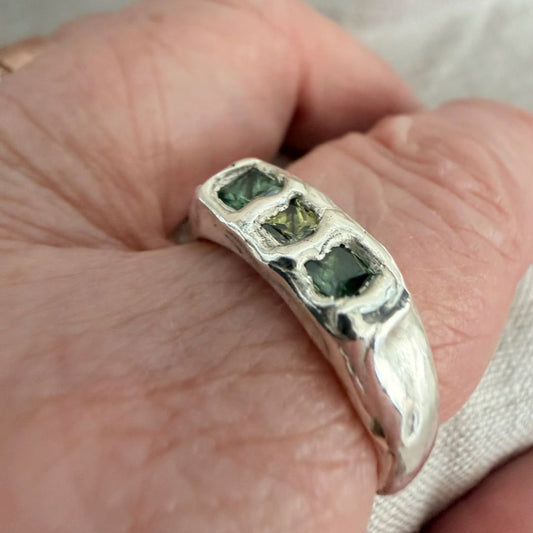 Kākāriki sterling silver signet ring worn on a finger, showing its softly polished organic band and three green princess-cut sapphires.