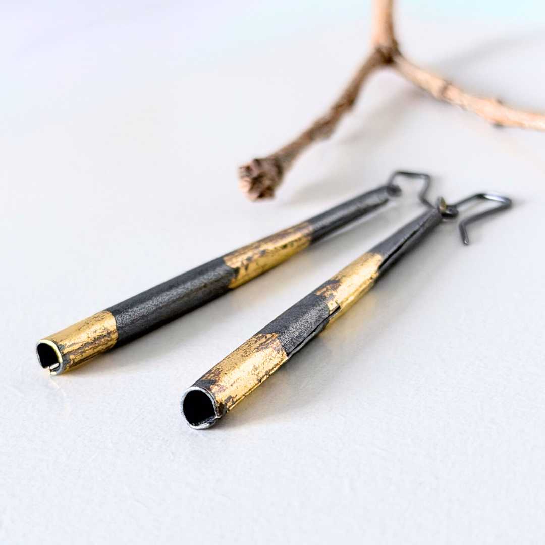 Keum-Boo pendant earrings with hand-applied gold detail resting beside a branch, photographed from a slight overhead angle.